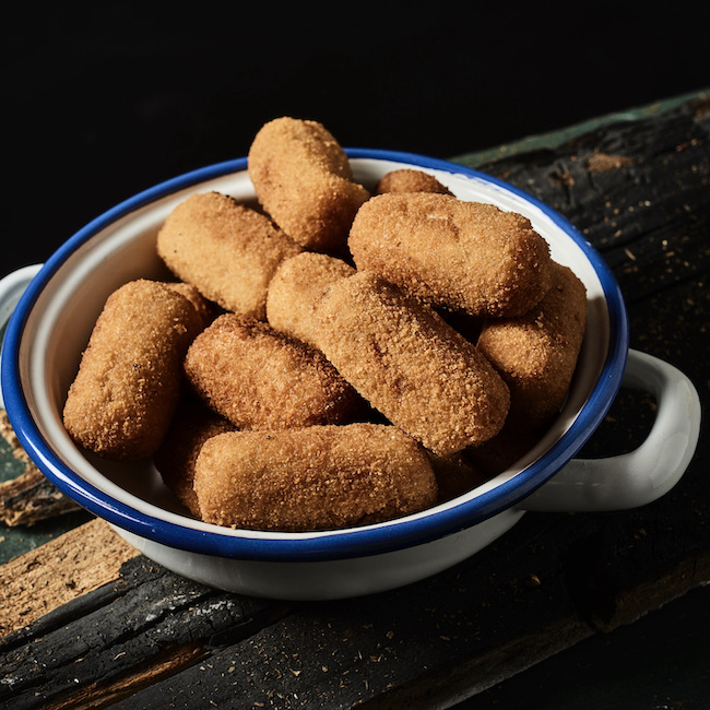 closeup of some spanish croquettes in a white and blue enamel plate on a rustic dark gray wooden table, against a black background