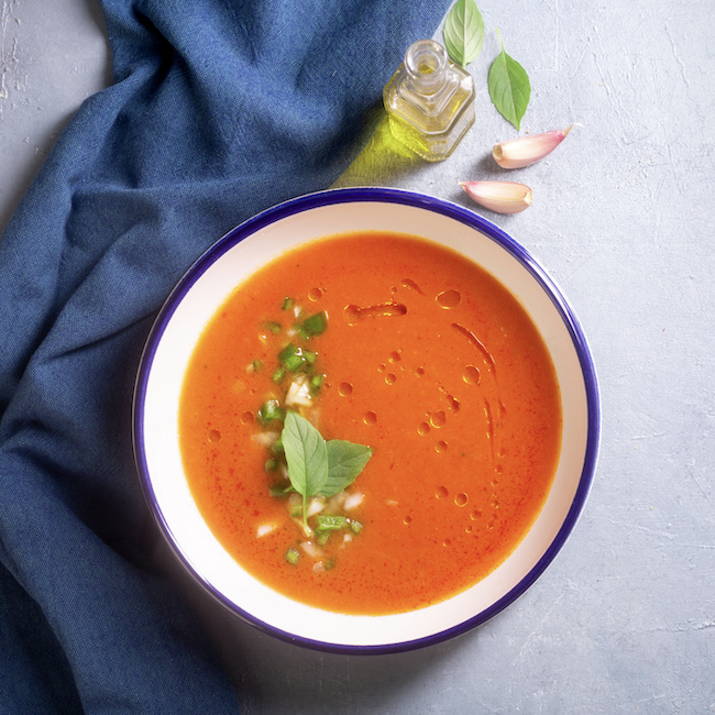 A plate of gazpacho made from tomato with garlic and basil leaves and olive oil on a gray background with a blue cloth copy space. Traditional Spanish dish