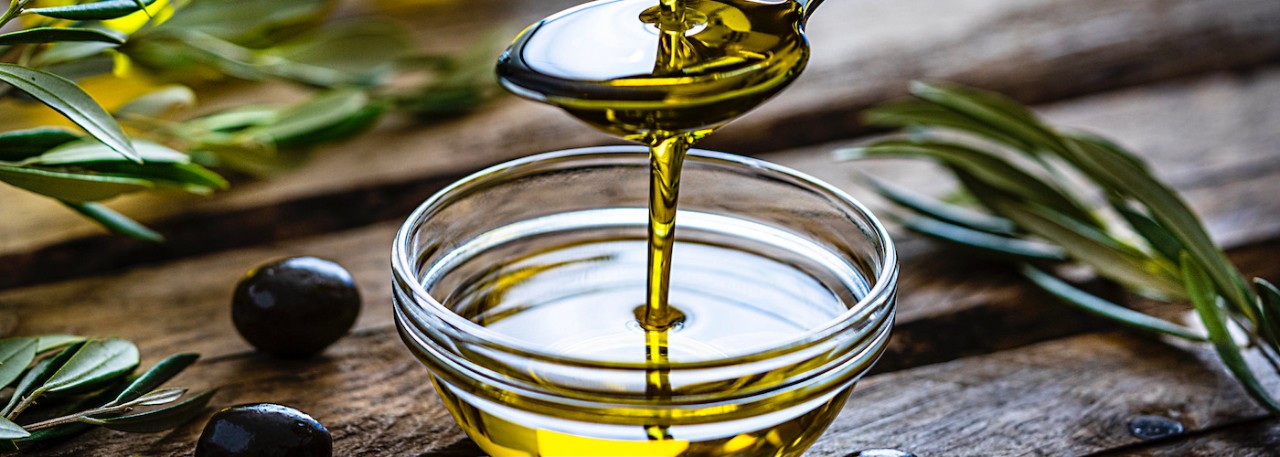 Pouring extra virgin olive oil from a spoon to a glass container. Olive branches comes from the left and right. Some olive oil bottles are out of focus at background. Black olives are on the table and complete the composition. The composition is on a rustic wooden kitchen table. Predominant colors are gold, green and brown. High resolution 42Mp studio digital capture taken with Sony A7rII and Sony FE 90mm f2.8 macro G OSS lens